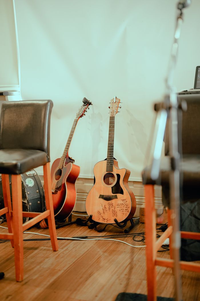 Home Two acoustic guitars resting indoors, surrounded by chairs, with a warm and inviting ambiance.