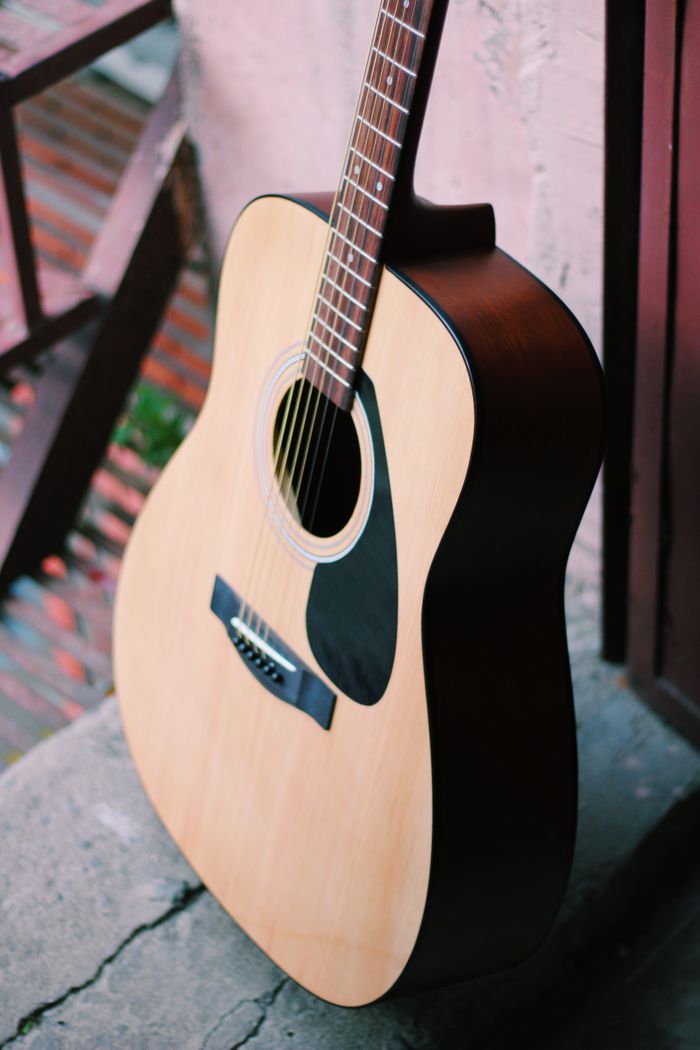 About An acoustic guitar with a light wooden finish leaning against an outdoor wall.